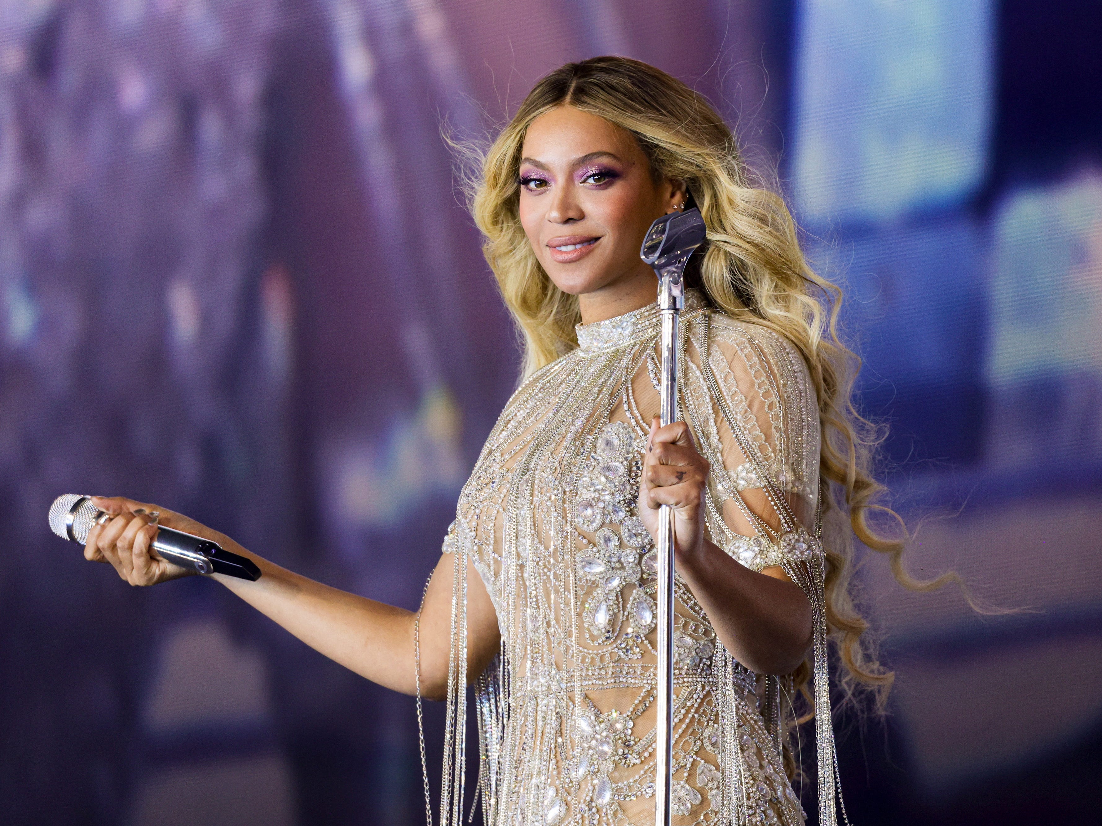 Beyoncé onstage during the Renaissance tour, holding a microphone out to the audience and wearing a diamond-encrusted outfit.