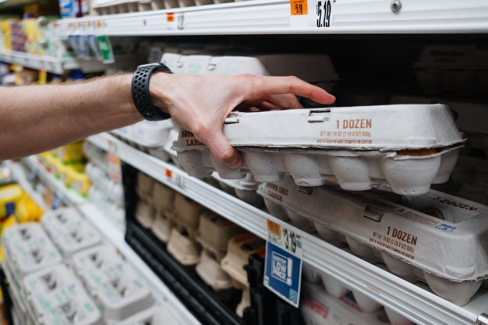 A stock image of a white man's hand grabbing a dozen eggs from a store shelf.