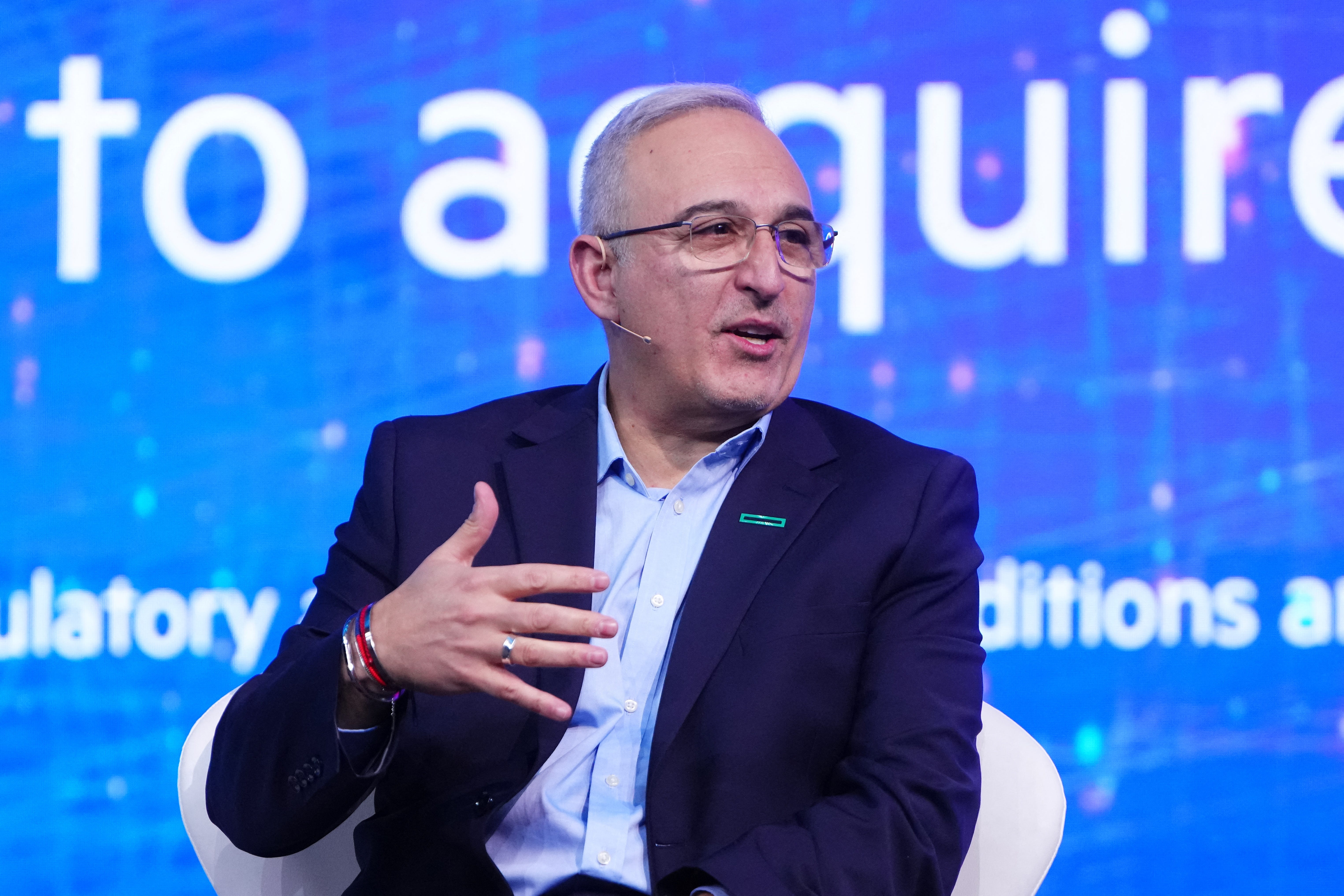 A man with grey hair wears a blue collared shirt and dark blue shirt. He gestures as he speaks while sitting on a stage in front of a large blue screen.